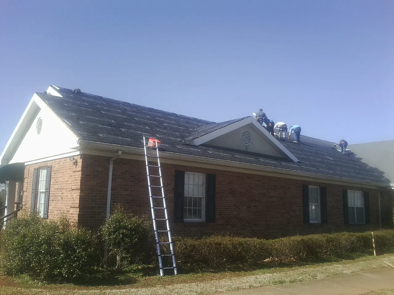 Crew tearing shingles off a brick ranch roof with a ladder set — Emergency Roof Repair work by our crew in Santa Claus