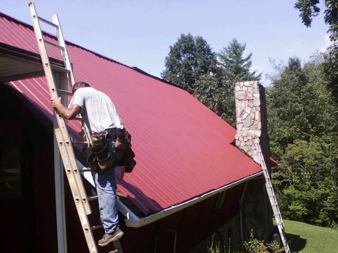 Roofer on a ladder beside a red metal roof and stone chimney — Soffit & Fascia Repair by Roofing Brothers in Santa Claus